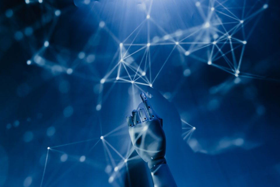Mão robótica alcançando uma rede digital iluminada em fundo azul, em representação conceitual da inteligência artificial e da tecnologia em rede